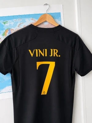 Real Madrid svart matchtröja Vini Jr. - Säljer en svart Real Madrid matchtröja från Adidas med VINI JR. och nummer 7 i gult på ryggen. Tröjan har klubbmärke, sponsorlogga och Champions League-märke på ärmen. Tillverkad i funktionsmaterial, perfekt för fotboll eller träning.
