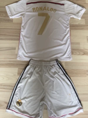 Real Madrid Ronaldo matchställ - Säljer ett vitt Real Madrid matchställ från Adidas med Ronaldo 7 på ryggen. Tröjan har klubbmärke, Fly Emirates-tryck och svarta detaljer på axlarna. Shortsen har klubbmärke och svarta ränder längs sidorna. Perfekt för fotbollsträning eller fans.
