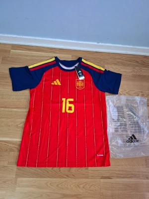 Spaniens matchtröja Rodrigo #16 Adidas VM 26/27 - Säljer en officiell röd Spanien matchtröja från Adidas med gula detaljer och nummer 16 samt namnet Rodrigo på ryggen. Tröjan har blå ärmar, gula ränder på axlarna och klubbmärke på bröstet. Perfekt för dig som älskar fotboll och vill stötta landslaget.