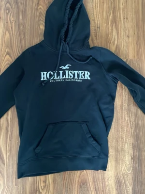 Svart hoodie från Hollister - Snygg svart hoodie från Hollister med stor logga och texten 'Southern California' på bröstet. Klassisk känguruficka framtill och justerbar huva med dragsko. Perfekt för en avslappnad och trendig look.