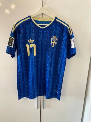 Blå Sverige fotbollströja Gyökeres #17 - Säljer en blå matchtröja från Sveriges landslag med Gyökeres och nummer 17 i gult på ryggen. Tröjan har gulddetaljer, officiella FIFA-märken på ärmarna och ett unikt mönster över hela tyget. Tillverkad i lätt och ventilerande material, perfekt för fotboll.