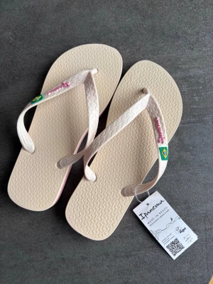 Beige flip-flops från Ipanema - Snygga beige flip-flops från Ipanema med vita remmar och rosa detaljer. Remmarna har en liten brasiliansk flagga och Ipanema-logga. Sulan är platt med rosa rand längs kanten och mönstrad ovansida. Perfekta för sommaren eller stranden.