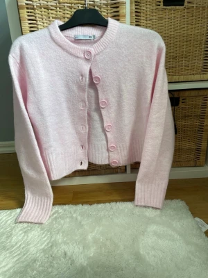 Ljusrosa fluffig kofta med knappar - Hely ny och oanvänd!! Supersöt ljusrosa kofta med rund hals och stora matchande knappar framtill. Koftan är fluffig och mjuk med ribbade muddar vid ärmslut och nederkant. Perfekt för dig som gillar en mysig och trendig look.