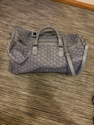 Grå weekendbag  - Snygg grå weekendbag med klassiskt mönster och detaljer i läder. Väskan har både handtag och justerbar axelrem samt dragkedja upptill. Perfekt för resor eller gymmet och rymmer allt du behöver. Stilren och tidlös design.