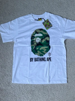 Vit BAPE t-shirt med camo-print - Säljer en vit t-shirt från BAPE med det klassiska kamouflageapansikte-trycket på bröstet och texten 'BY BATHING APE' under. T-shirten har rund hals, korta ärmar och är gjord i mjuk bomull. Märkeslapp med BAPE-logga på ärmen.