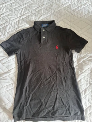 Svart piké från Polo Ralph Lauren - Klassisk svart pikétröja från Polo Ralph Lauren i slim fit-modell. Tröjan har korta ärmar, krage med två knappar och den ikoniska röda logotypen broderad på bröstet. Tillverkad i mjuk bomull som känns skön mot huden.