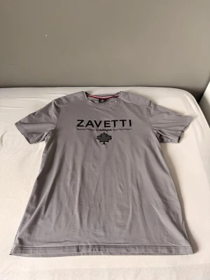 Grå t-shirt från Zavetti Canada - Snygg grå t-shirt från Zavetti Canada med svart logga och lönnlöv på bröstet. Klassisk rund halsringning med en cool detalj i form av röd och svart rand på insidan av kragen. Perfekt till jeans eller shorts.