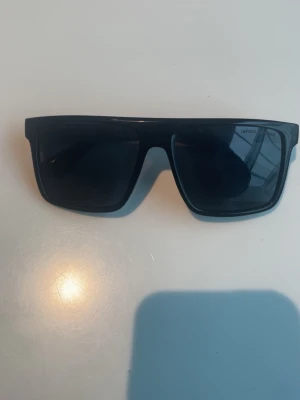 Svarta solglasögon Emporio Armani - Snygga svarta solglasögon från Emporio Armani med breda bågar och rektangulär form. Glasögonen har en modern och stilren look med mörka glas och diskret logga på sidan. Perfekta för att höja din outfit till nästa nivå.