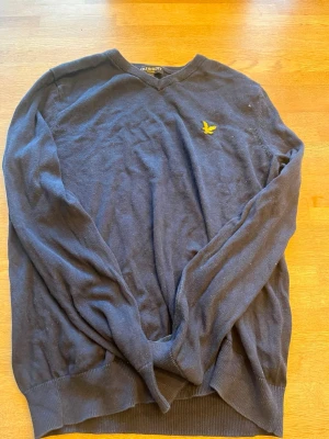 Mörkblå stickad tröja Lyle & Scott - Mörkblå stickad tröja från Lyle & Scott med V-ringning och den klassiska gula logon broderad på bröstet. Tröjan har långa ärmar och ribbade muddar vid ärmslut och nederkant. Perfekt för en stilren och avslappnad look.