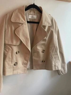Kort beige trenchcoat från H&M - Snygg kort trenchcoat från H&M i beige bomullsmix. Jackan har dubbelknäppning med stora knappar, breda slag och två sidofickor. Perfekt för dig som gillar klassisk stil med en modern twist. Storlek 152 men jag rekommenderar storlek XS👌