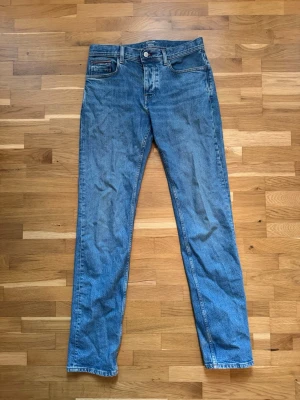 Blå raka jeans från Tommy Hilfiger - Klassiska blå jeans från Tommy Hilfiger med rak passform och fem fickor. Jeansen har en enkel design med logotypdetalj på bakfickan och patch bak i midjan. Tillverkade i denim, perfekta för en avslappnad stil.