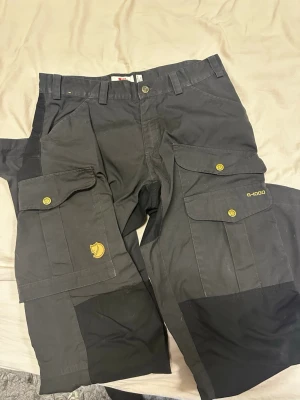 Svarta cargobyxor Fjällräven G-1000 - Säljer ett par svarta cargobyxor från Fjällräven i slitstarkt G-1000-material. Byxorna har flera stora fickor med knappar, förstärkta knän och detaljer i gult. Perfekta för dig som gillar funktionella och tåliga byxor med cool outdoor-vibe.  Ny pris:2300