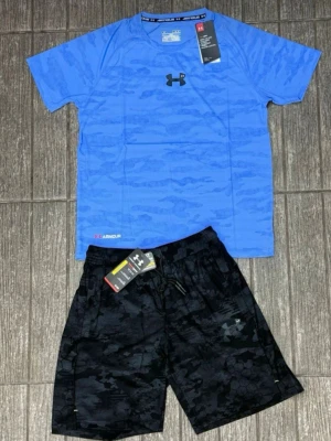 Blå träningsset från Under Armour - Snyggt träningsset från Under Armour med en blå t-shirt i camomönster och svarta shorts med diskret mönster. T-shirten har rund hals och logga på bröstet, shortsen har dragsko i midjan och sidofickor. Perfekt för gym eller löpning.