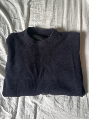 Mörkblå stickad tröja från Jack & Jones - En stilren mörkblå stickad tröja med rund halsringning och ribbade muddar. Tröjan har en klassisk passform och är tillverkad i ett mjukt, tättstickat material som passar perfekt till kyligare dagar men även till sena sommarkvällar.