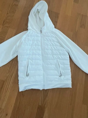 Vit pufferjacka med huva  - Snygg vit pufferjacka  med dragkedja framtill och två fickor med dragkedja . Jackan har en quiltad framsida och mjuka ärmar samt en stor huva. Perfekt för kalla dagar och enkel att matcha med olika outfits. Passar xs och s