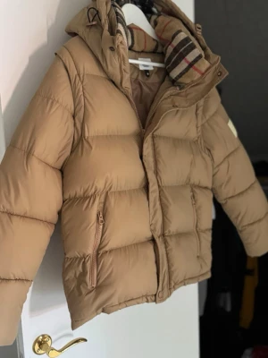Beige pufferjacka från Burberry - Snygg beige pufferjacka från Burberry med klassiskt rutigt foder i huvan. Jackan har dragkedja framtill, två sidofickor med dragkedja och en hög krage. Perfekt för kalla vinterdagar och riktigt trendig look.