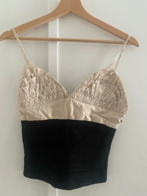 Beige och svart topp med tunna band - Snygg topp med tunna axelband och tvådelad design i beige och svart. Överdelen har rynkade kupor och smockad midja för en tight passform. Perfekt att styla med jeans eller kjol för en trendig look.
