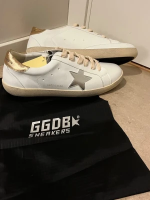 Golden Goose Skor - Golden Goose sneakers i vitt läder med metallicguld på hälen och en silverfärgad stjärna på sidan. Beige skosnören och GGDB-tryck på sidan. Klassisk låg modell med slitstark sula och snygg kontrast mellan färgerna. Perfekta för en trendig streetstyle-look.