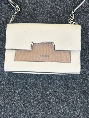 Vit och beige kuvertväska Calvin Klein - Snygg kuvertväska från Calvin Klein i vitt och beige konstläder. Väskan har en stilren design med silverfärgad kedjerem och logga framtill. Perfekt för dig som gillar minimalistisk och modern stil.