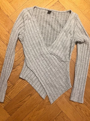 Vår Grå ribbad omlottopp - Säljer en grå ribbad omlottopp med lång ärm och djup v-ringning. Toppen har en asymmetrisk nederkant och är tillverkad i mjukt stretchigt material. Perfekt för dig som gillar en stilren men ändå unik look.