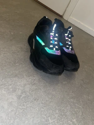 svarta sneakers - Säljer ett par chunky svarta sneakers med reflekterande detaljer och meshpaneler. Skorna har en kraftig sula och coola färgskiftande partier som syns i ljus. Perfekta för dig som vill sticka ut med din stil.