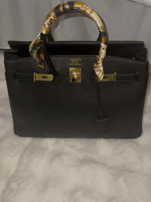 Svart handväska 35 cm - Säljer en svart Birkin-liknande handväska. Icke använd då den var för stor för min smak. Kommer med plast innuti för att hålla formen samt silk sjalen som man kan ta bort om man önskar. 