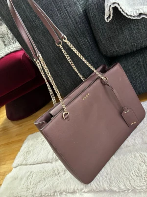 Brun handväska från DKNY med kedjerem - Brun handväska från DKNY i en rektangulär modell. Bra skick! Väskan har dubbla axelremmar i läder och kedja, samt en liten tag med DKNY-logga. Tillverkad i ett slätt, tåligt material med guldfärgade detaljer. Fickor inuti, 2 med dragkedja. Pris kan diskuteras.