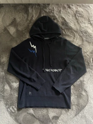 Svart hoodie från Monalto - Svart hoodie från Monalto med huva och dragsko. Framtill finns vita och blå blixtdetaljer samt texten 'C'EST MONALTO'. På ryggen står det 'MADE IN FRANCE' i vit text. Hoodien har en avslappnad passform och är tillverkad i mjukt material.