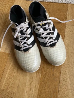 Adidas vita och svarta innetränings skor  - Säljer ett par Adidas fotbollsskor i vitt, svart och guld. Skorna har klassiska tre ränder på sidan, snörning och ovandel i syntetmaterial med textur för bättre bollkontroll. Grymma för dig som vill ha stil i fotsal 