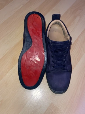 Mörkblå sneakers med röd sula - Snygga mörkblå sneakers med blå snörning och kontrasterande röd sula. Skorna har en klassisk låg profil och är tillverkade i mockaliknande material med beige innerfoder. Perfekta för dig som vill ha en stilren och unik look.