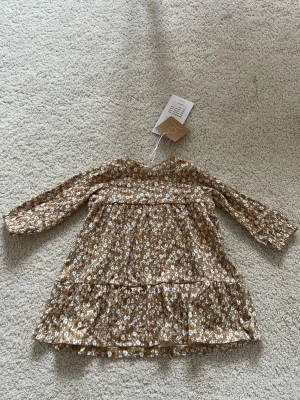 Blommig beige klänning från Name It - Söt beige klänning från Name It med små vita blommor och långa ärmar. Tillverkad i mjuk ekologisk bomull, perfekt för vår och sommar. Klänningen har en avslappnad passform och volang nedtill för extra rörelsefrihet.