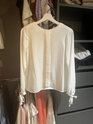 Vit blus med knytband vid ärmar - Stilren vit blus med rund halsringning och långa ärmar som avslutas med dekorativa knytband vid handlederna. Blusen har en rak passform och knappdetalj i nacken. Perfekt för en clean och enkel look.