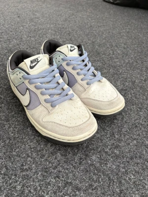 Nike Dunk Low i grå och ljusblå - Snygga Nike Dunk Low sneakers i grått och ljusblått med klassisk swoosh på sidan. Skorna har perforerad tåbox, ljusblå skosnören och vadderad häl. Ovandelen är i mocka och textil, och insidan är fodrad i svart tyg.