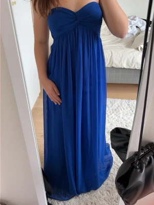 Blå axelbandslös maxiklänning - Snygg blå axelbandslös maxiklänning med draperad överdel och lätt utställd kjol. Klänningen har en romantisk och elegant look, perfekt för speciella tillfällen. Tillverkad i ett tunt, flygigt material som ger ett fint fall. perfekt för speciella tillfällen som balen, bröllop och studenten. Storlek 34 känns som XS