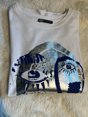 Vit t-shirt med konstnärligt tryck Zara - Vit t-shirt från Zara med ett stort konstnärligt ansiktstryck i blått och metallic silver på framsidan. T-shirten har rund hals och korta ärmar. Materialet är mjuk bomull som känns skönt mot huden. Använd max 3 gånger.