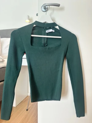 Mörkgrön topp med cut out från Zara - Trendig mörkgrön topp från Zara med lång ärm och snygg cut out-detalj vid halsen. Toppen har en tight passform och dragkedja bak. Perfekt för dig som vill ha en stilren men unik look.