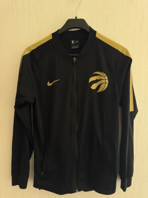 Svart Raptors jacka från Nike - Säljer en svart zip-jacka från Nike med Toronto Raptors logga och guldfärgade detaljer. Tillverkad i ett sportigt material. Pris kan diskuteras