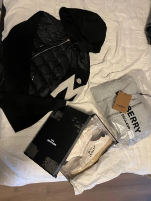 Märkes kläder endast budning - Säljer en svart hoodie från Moncler med dragkedja och vadderade partier framtill. Tröjan har en stor huva och ett vitt zigzag-mönster på ena ärmen samt Moncler-logga på bröstet. Perfekt för dig som gillar streetwear och exklusiva märken.