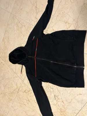 Svart hoodie från  Hugo boss med dragkedja - Snygg svart hoodie från  Hugo boss  med dragkedja framtill och röda detaljer över bröstet. Klassisk huva med dragsko och två fickor på sidorna. Tillverkad i bomullsmix för en bekväm känsla. Perfekt för en avslappnad streetwear-look.