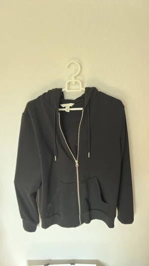 Svart hoodie med dragkedja från H&M - Klassisk svart hoodie från H&M med dragkedja framtill och två fickor. Tröjan har huva med dragsko och ribbade muddar vid ärmslut och nederkant. Perfekt för en avslappnad och stilren look. Tillverkad i mjuk bomullsblandning. Skriv gärna pris förslag!