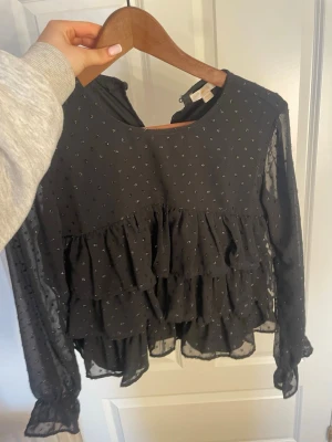 Svart volangblus med prickar - Svart blus från Gina Tricot med transparenta långärmar och små silverprickar. Blusen har flera volanglager framtill och är gjord i ett tunt, luftigt material som ger en cool och trendig look. Rund halsringning och knäppning bak i nacken. Bra skick, använd ca 3 gånger. Kan skicka fler bilder, lägre pris vid direkt köp