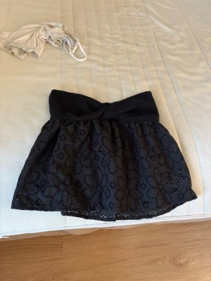 Svart kortkjol med broderade blommor - Supersöt svart kortkjol med bred ribbad midja och broderade blommor i tyget. Kjolen har en omlottliknande midjedetalj och är perfekt för dig som gillar en feminin och trendig stil.