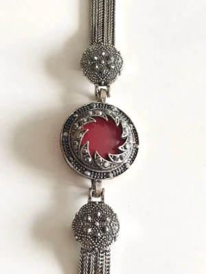 Silverfärgat armband med röd sten - Unikt armband i silverfärgad metall med flera kedjor och detaljerade pärlprydda dekorationer. I mitten sitter en rund röd sten omgiven av ett snirkligt mönster. Armbandet har vintagekänsla och cool design som sticker ut.
