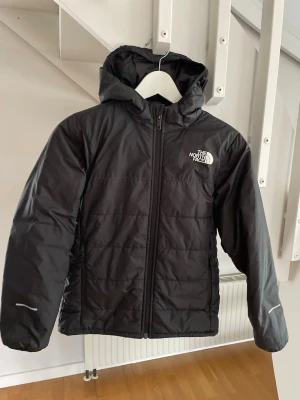 Svart pufferjacka från The North Face - Säljer en svart pufferjacka från The North Face med huva och dragkedja framtill. Jackan har diskret logga på bröstet och ryggen, samt reflekterande detaljer på ärmarna. Tillverkad i slitstarkt material som håller värmen under kalla vinterdagar.