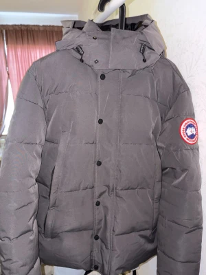 Grå dunjacka från Canada Goose - Snygg grå dunjacka från Canada Goose med klassisk patch på ärmen. Jackan har huva, raka linjer och stora paneler för extra värme. Perfekt för kalla dagar och har en modern, stilren look med knappar framtill.