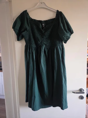 Grön smockad klänning med puffärm, stl XL - Mörkgrön klänning med smockad överdel och puffärmar. Klänningen har en v-ringad halsringning och är tillverkad i ett mjukt bomullstyg. Kjolen är vid och faller fint till under knäet. Perfekt för dig som gillar en romantisk och avslappnad stil.
