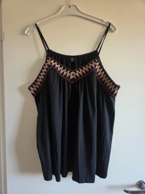 Svart linnetopp med färgglad kant - Säljer en svart linnetopp med tunna axelband och lös passform. Toppen har en färgglad, sicksackmönstrad kant längs ringningen som ger en unik touch. Perfekt för varma dagar och enkel att styla med jeans eller kjol.