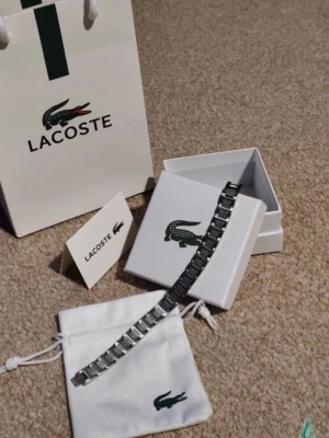 Silverfärgat armband från Lacoste - Helt ny oanvänd snyggt silverfärgat armband från Lacoste med länkdesign och diskreta detaljer. Armbandet är gjort i metall och har en stilren, modern look med Lacoste-loggan på förpackningen. Perfekt accessoar för dig som gillar klassisk men trendig stil.ett till ett, fraktar snabbt:)
