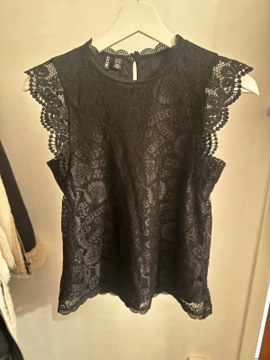 Svart spetsblus från Vero Moda  - Supersnygg svart blus i spets från Vero Moda med fina detaljer och mönster. Ärmlös modell med dekorativa spetskanter vid axlarna och halsringning. Perfekt att styla med jeans eller kjol för en trendig look.