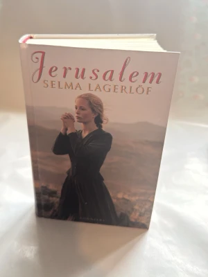 Jerusalem av Selma Lagerlöf - Säljer romanen 'Jerusalem' av Selma Lagerlöf i en snygg inbunden utgåva från Bonnier. Omslaget har en mjuk, matt finish med ett foto av en kvinna mot ett landskap i jordnära toner. Perfekt för dig som gillar klassisk svensk litteratur.